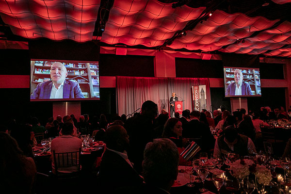 A large banquet hall filled with people seated at round tables looking up at two large screens showing Gregory Gatta addressing them.