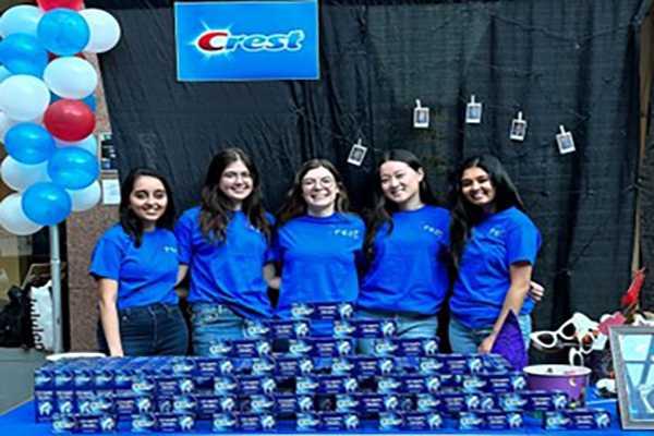 Image of Jenny Hamilton standing, smiling with four other classmates in blue t-shirts and jeans. They stand under a “Crest” logo banner at a blue booth for Johnson’s Battle of the Brands competition.