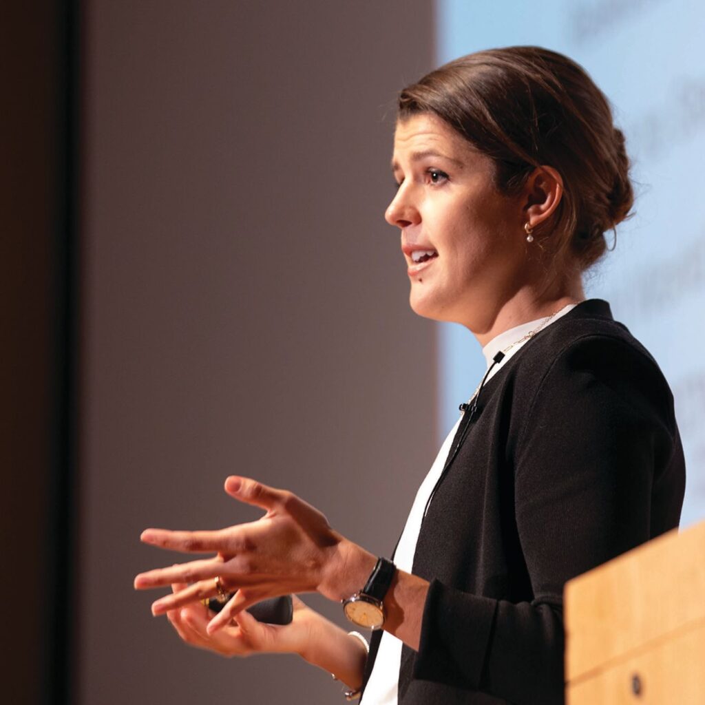 Heggie speaks next to a podium in font of a projector screen.