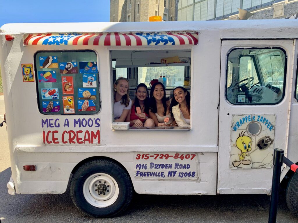 Four women smile from within the service window of a white ice cream truck.