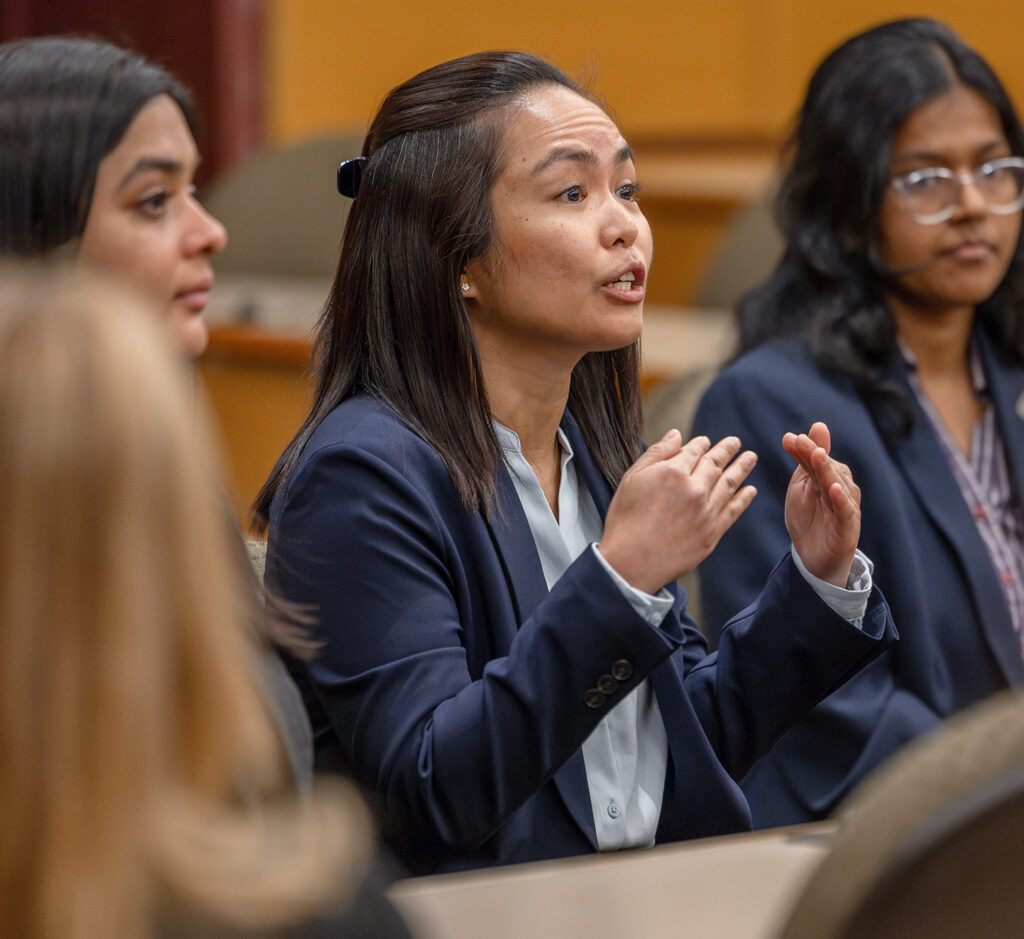 A female student in business attire gestures while speaking, with other students visible in the foreground and background.