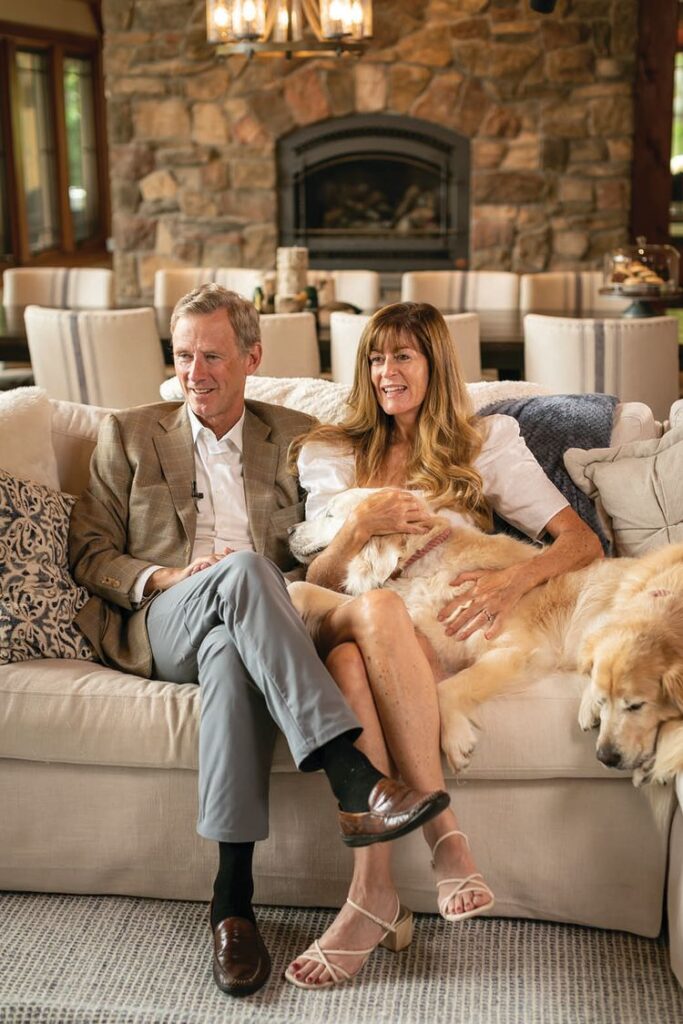 Peter and Stepanie Nolan sit on their couch with one dogs head on Stephanie's lap and another laying on the couch next to her.