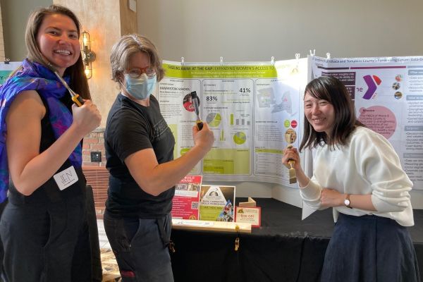 Jennifer White-Phalen and Miwako Nara Uemura stand in front of their project’s poster. Hammerstone founder Maria Klemperer-Johnson has carpentry tools in hand.