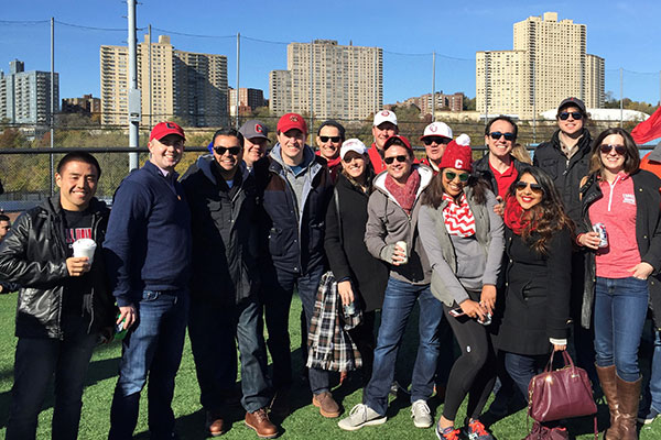 A group photo of 15 people, many wearing red Cornell hats, with tall buildings and blue sky in the background.