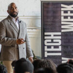 A man standing in front of a room full of people next to a Tech Week banner and speaking.