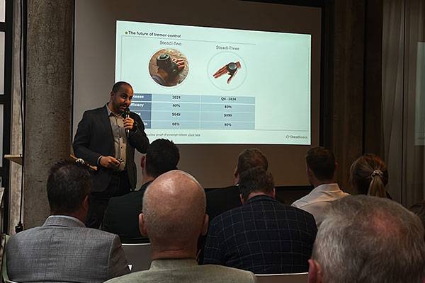 A man standing on stage and sepaking into a microphone next to a screen showing Steadiwear's product and some product information.