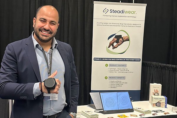 A man wearing a round device attached to the back of his hand and standing next to a table and poster displaying information about Steadiwear.