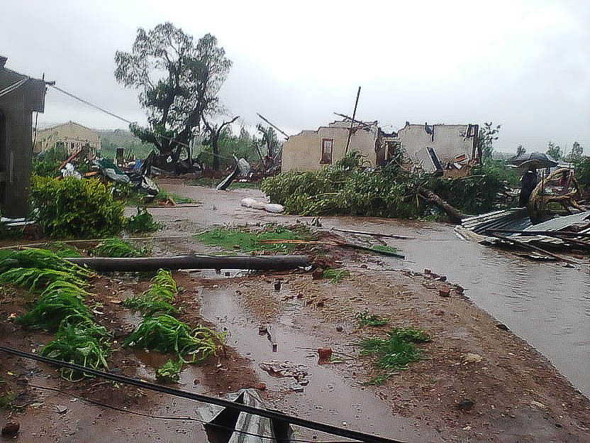 Cyclone-damaged buildings and latrines lack roofing, and debris is strewn across muddy flooded roads.