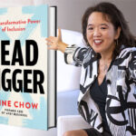A woman in a black and white blazer with her hands outstretched to the right of an enlarged image of the book she authored.