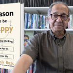 Man stands in front of a book shelf and to his left is an enlarged image of the book he authored.