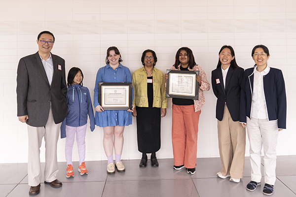The Ye Family, father, mother and two young daughters, smile for a posed photo with the associate director of graduate programming and two female graduate students who are holding their award certificates
