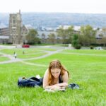 A student lays on her belly reading on the grass, with Cornell's West Campus in the background.