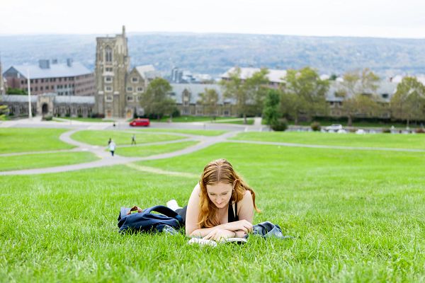 A student lays on her belly reading on the grass, with Cornell's West Campus in the background.
