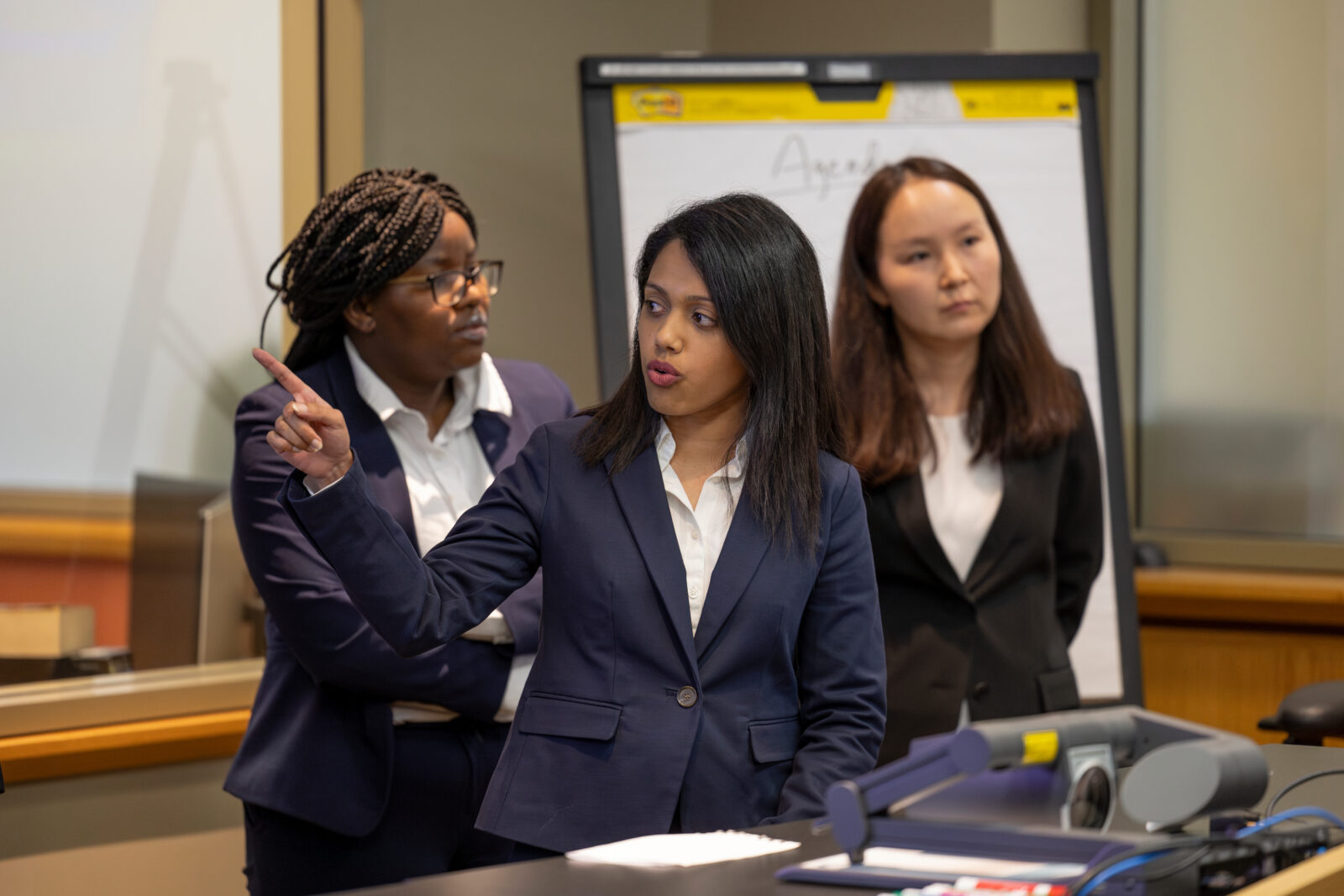 Photo of three woman standing behind a desk, presenting to an off-screen audience. One woman in the front is pointing to a screen off camera.