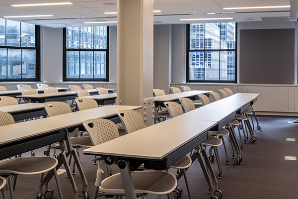 Rows of classroom tables and chairs with NYC buildings visible out the windows.