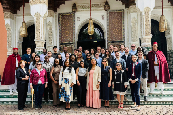A large group of people dressed in formal, semiformal, and traditional attire stands on the steps of an ornate building with arched doorways, decorative columns, and hanging lanterns