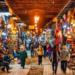 A bustling market street lined with colorful shops and stalls displays textiles, bags, and lanterns, with people walking through under a lattice-covered ceiling illuminated by hanging lanterns.