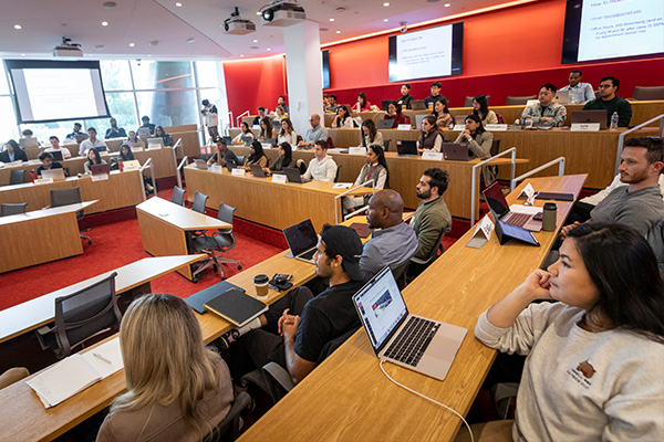 Students sit at tiered classroom tables with a wall of windows visible behind a projector screen.