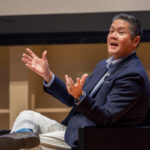 A man sits on stage talking with his hands outstretched.