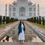 Vanessa stands in front of the Taj Mahal, which is reflected in a long rectangular pool beneath a sky dotted with clouds.