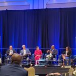 Panel of seven speakers seated on stage with banners.
