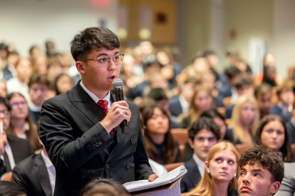 A student wearing a suit stands with a microphone in a full auditorium.