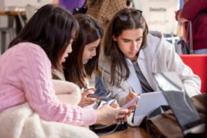 Three women hunch over a laptop and phones.