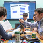 Five students work at a table with a large screen on the back wall of the room.