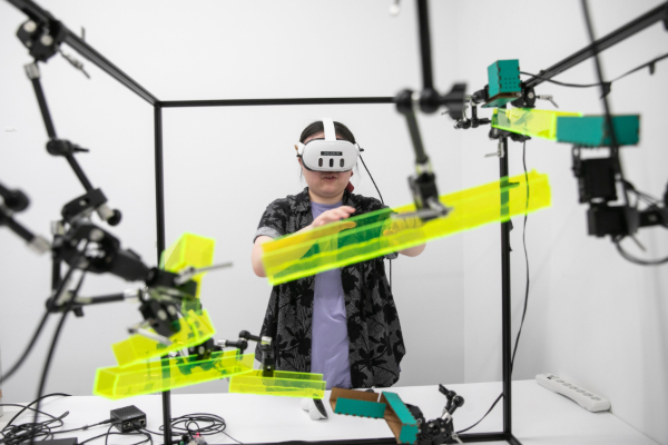 A student wearing VR goggles stands behind equipment. 