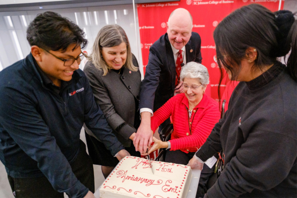 Two people hold a big cake while three others hold the knife, poised to cut.