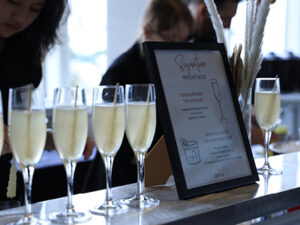 A row of light yellow cocktails on a counter top next to a menu card listing its ingredients.