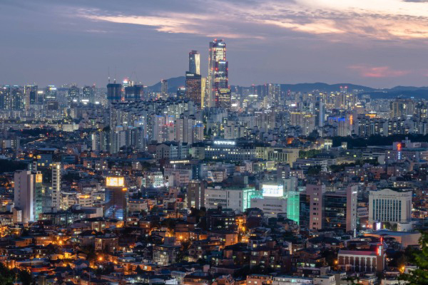 A wide view of Seoul, South Korea, at dusk shows dense low-rise neighborhoods in the foreground and brightly lit modern skyscrapers rising in the distance