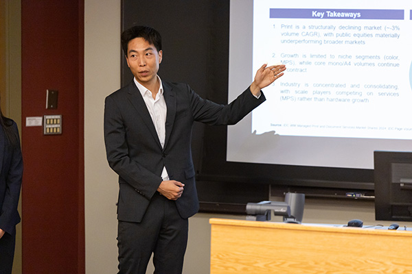 Male student standing in front of a classroom, speaking to the class and pointing at the screen behind him.