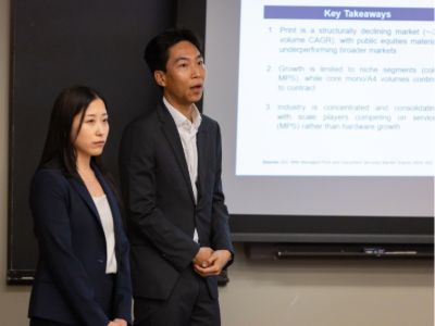 Two students standing in front of a screen, speaking to a class outside of the shot of the photo.