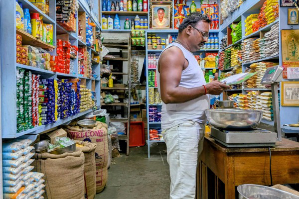A person stands at a counter inside a small, densely stocked grocery, reading a package beside a metal scale, with shelves of colorful packaged goods and sacks of grains lining the narrow aisles