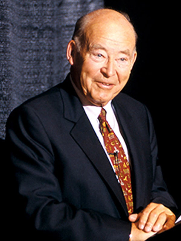 Samuel Curtis Johnson speaking during a panel discussion at Cornell, wearing a suit and tie against a dark curtain backdrop.