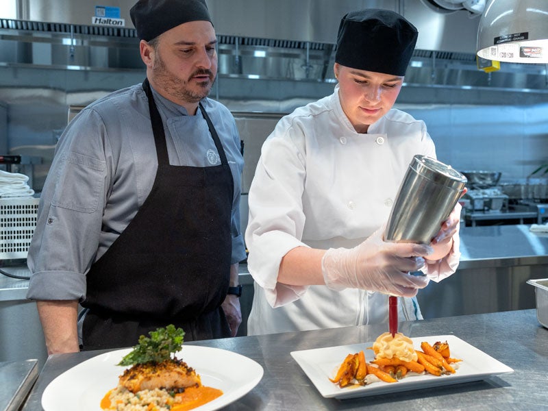 A student chef at Cornell University&rsquo;s Nolan School plates a dish in a professional kitchen as an instructor observes.