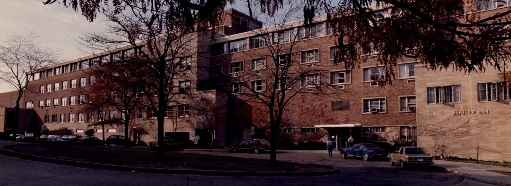 A photo of Statler Inn with cars parked in front of it and trees in the foreground.