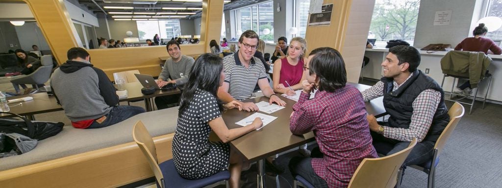 Open study lounge with wood pod seating and tables, with bright windows, modern chairs, and spaces for small groups.