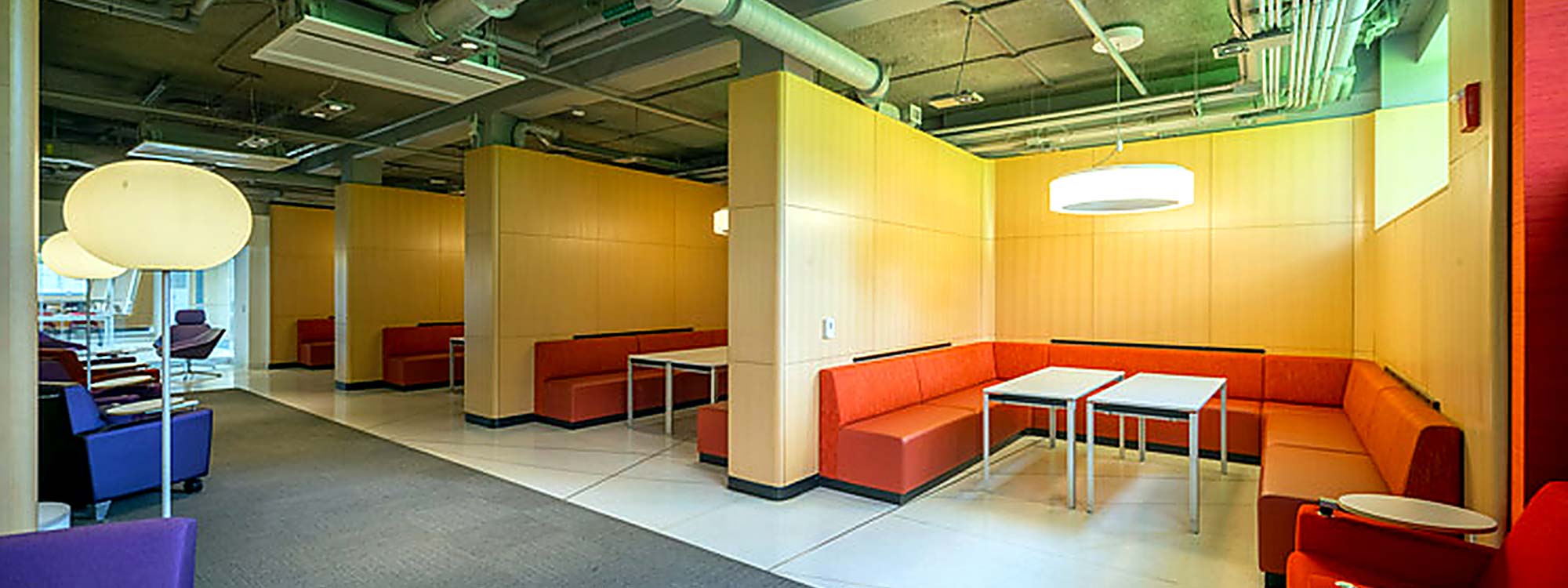 Modern lounge with open seating areas along a wide walkway. Orange upholstered benches and small white tables, divided by wooden panels.