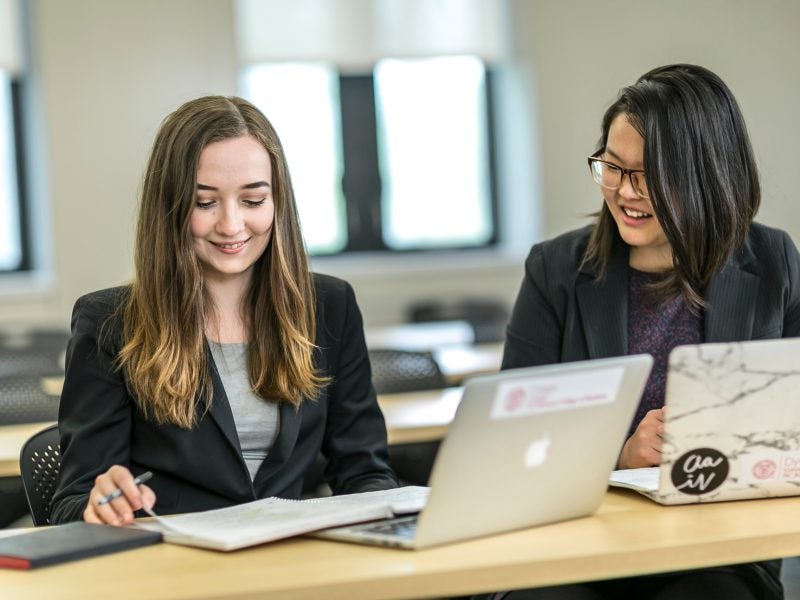 Two students sit at a table with their laptops