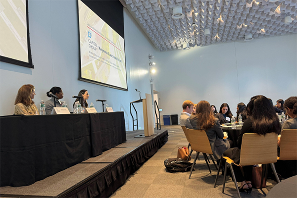Students sitting at round tables, looking at a stage with three women speaking on a panel