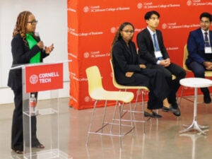 Fridah speaking on stage, with her students and partners seated in the background, listening to the presentation. A red Cornell banner is visible in the background.