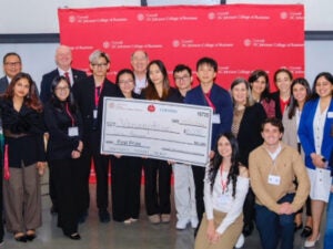 Finalists standing and crouching for a photo, with winners holding the check, surrounded by organizers and speakers. A red Cornell banner is in the background.