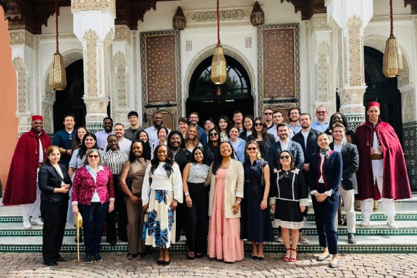 A large group of people dressed in formal, semiformal, and traditional attire stands on the steps of an ornate building with arched doorways, decorative columns, and hanging lanterns