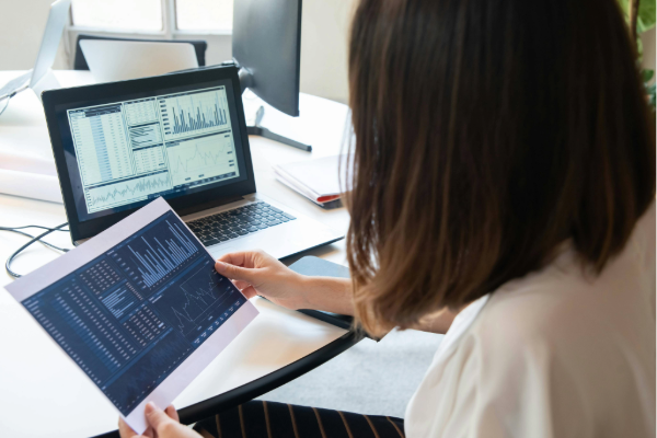 A Woman holding a paper with charts near her laptop.