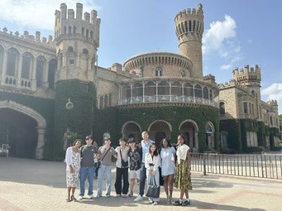 A group of people stands in front of Bangalore Palace, a historic castle-like building with ivy-covered walls and multiple towers, under a partly cloudy sky.