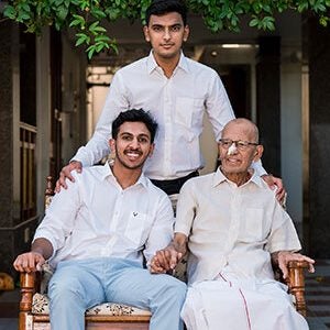 A young man and a much older man sitting on an upholstered wooden bench with a young man standing behind them, his hands on each shoulder. They are outside in a courtyard with foliage from a tree hanging over them.