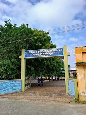 The entrance to school grounds with gates wide open, a sign overhead with writing in Tamil script, and students walking beyond the gate.