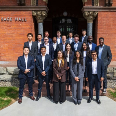 Group of 19 Cornell students dressed in business attire standing in front of Sage Hall on campus.